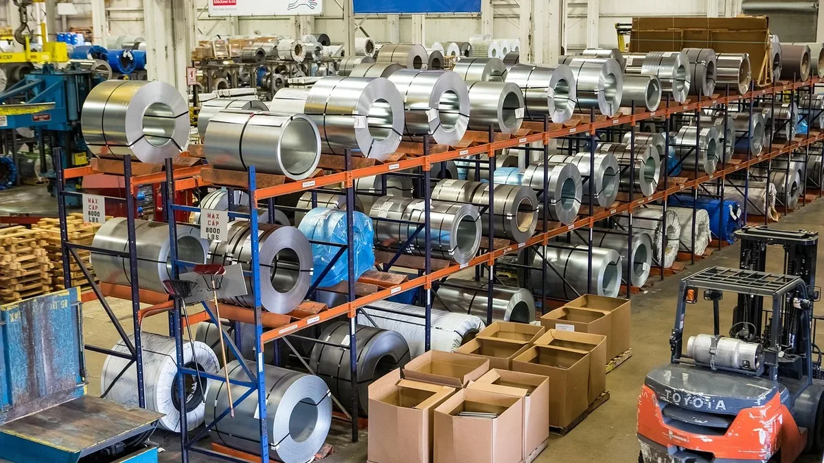 A warehouse or factory setting, featuring rows of shelving units stocked with large rolls of metal, cardboard boxes, and a forklift.