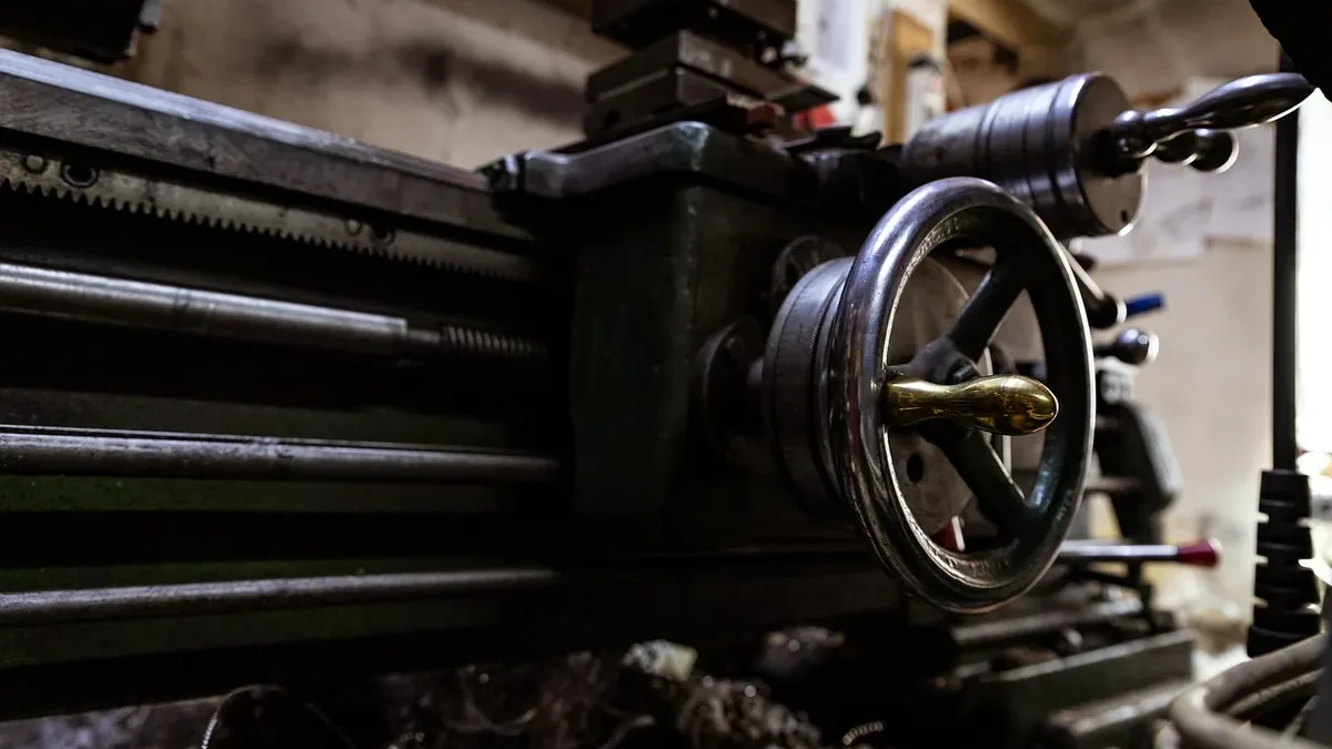 A large metal lathe with a wheel and handle, set against a backdrop of a workshop or factory.