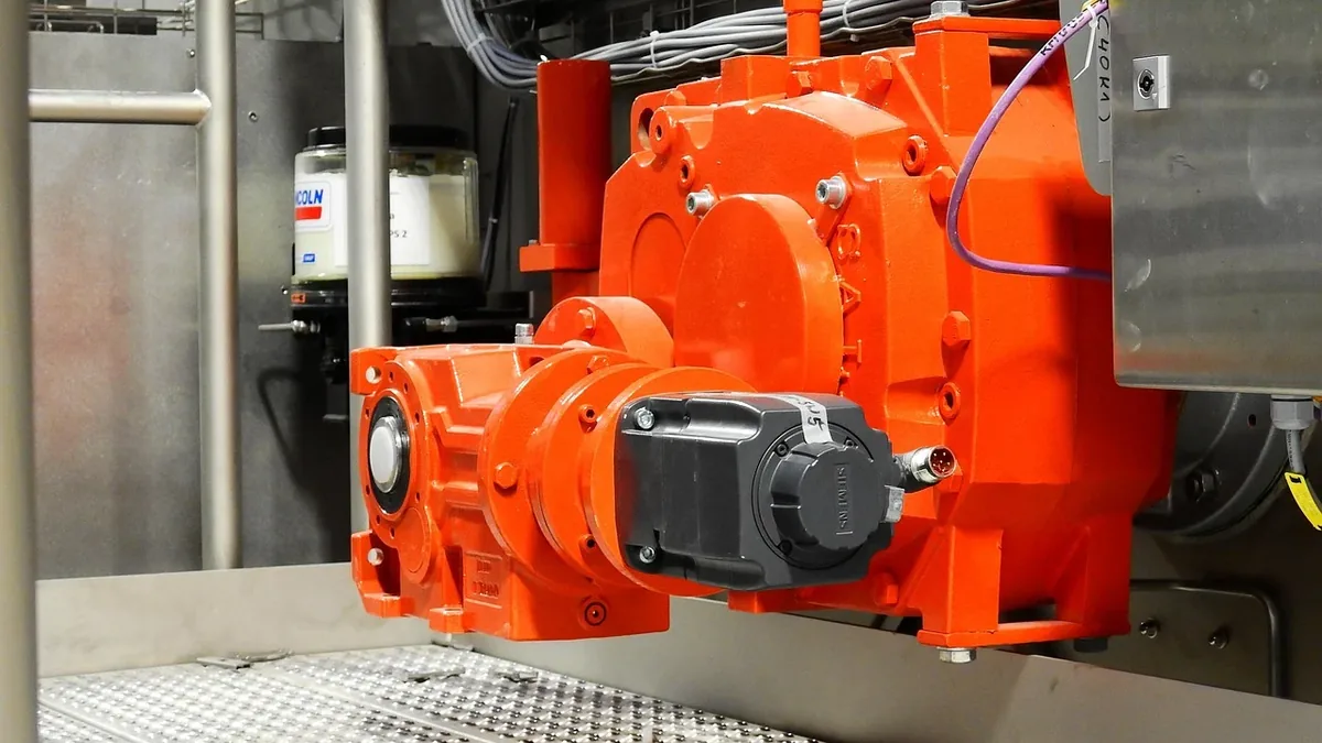 An industrial setting with a large orange machine featuring a black control panel and wires, situated on a metal grate.